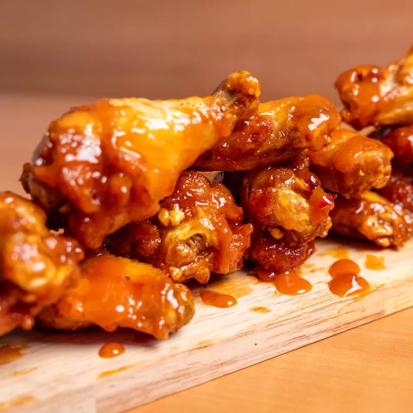 A close-up of several golden-brown chicken wings coated in a glistening orange sauce on a wooden board.