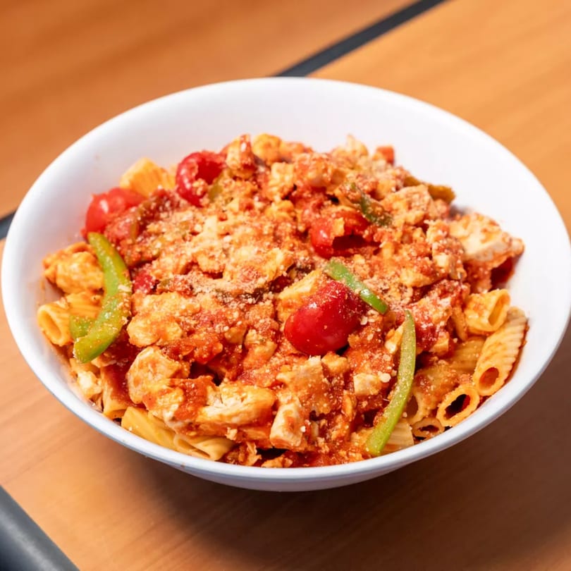 A white bowl of Capones Italian Eatery West pasta with red sauce, chicken, tomatoes, and green peppers.