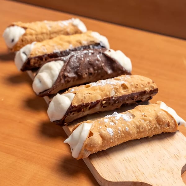 Five cannolis, some chocolate-dipped, filled with white cream and dusted with powdered sugar on a wooden board.