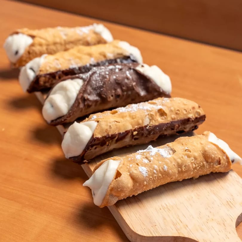 Five cannolis, some chocolate-dipped, filled with white cream and dusted with powdered sugar on a wooden board.